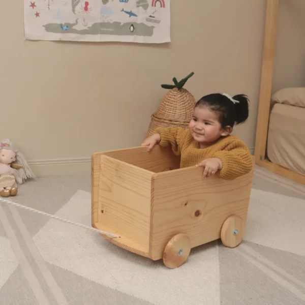 Niña usando carrito de arrastre de madera tipo caja con ruedas que permite transportar objetos y fomentar la autonomía en ambiente Montessori