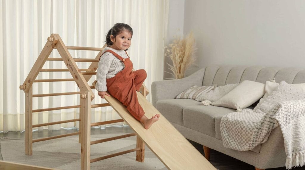 Niña pequeña usando triángulo Pikler Montessori con rampa para desarrollar equilibrio y movimiento en casa