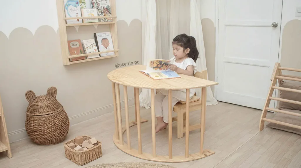 Niña sentada en mesa infantil Montessori de madera leyendo libro en ambiente preparado que fomenta autonomía y concentración