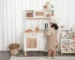 Niña pequeña jugando en una cocina Montessori de madera con fregadero, utensilios y estantería de libros infantiles accesible.